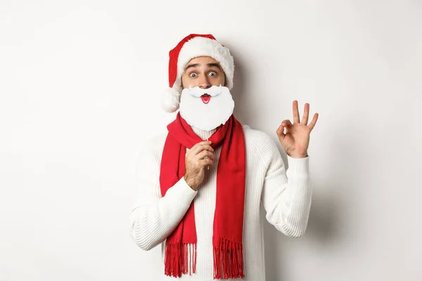 Festa de Natal e conceito de celebração. Modelo masculino feliz em chapéu de Papai Noel e máscara de barba branca, mostrando gesto ok, de pé sobre fundo branco — Fotografia de Stock