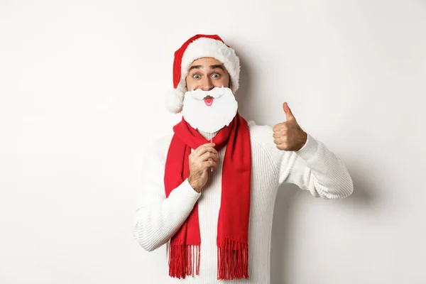 Festa de Natal e conceito de celebração. modelo masculino feliz em chapéu de Papai Noel e máscara de barba branca, mostrando polegar para cima gesto, de pé sobre fundo branco — Fotografia de Stock