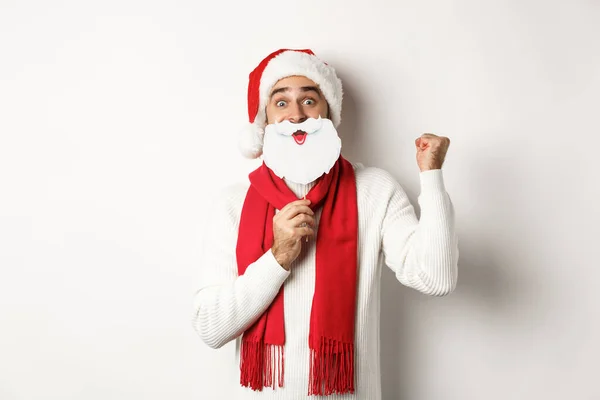 Festa de Natal e conceito de celebração. Jovem animado desfrutando de Ano Novo, olhando engraçado em chapéu de Santa com máscara de barba branca, fazendo punho bomba e regozijo — Fotografia de Stock