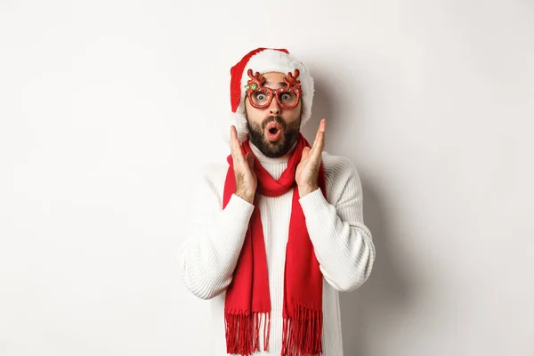 Рождество, Новый год и концепция празднования. Мужчина в очках для вечеринок и шляпе Санты выглядит удивлённым, слышит предложение о покупках, стоит на белом фоне — стоковое фото
