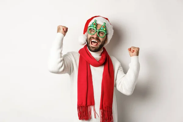 Noël, Nouvel An et concept de célébration. Joyeux modèle masculin dans les lunettes de fête et santa chapeau réjouissant, faisant poing pompe et triomphe, debout sur fond blanc — Photo