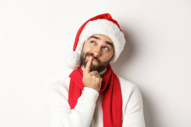 Yeni yıl partisi ve kış tatili konsepti. Noel Baba şapkalı sakallı adamın yakın çekimi sol üst köşede düşünceli görünüyor, düşünüyor ya da karar veriyor, noel hediyesini seçiyor, beyaz bir geçmişi var.