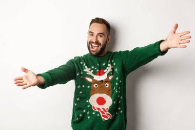 Yeni yıl kutlaması ve kış tatili konsepti. Arkadaş canlısı adam kucaklaşmak için ellerini açtı, Noel partisine hoş geldin, komik süveter giyiyor, beyaz arka plan
