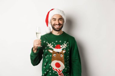 Noel partisi ve tatil konsepti. Noel Baba şapkalı ve komik kazaklı yakışıklı adam, şampanya içiyor ve yeni yılı kutluyor, beyaz bir geçmişi var.