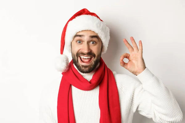 Neujahrsparty und Winterferienkonzept. Nahaufnahme eines glücklichen attraktiven Mannes mit Weihnachtsmannmütze, der Ok-Zeichen zeigt, Weihnachten feiert, weißer Hintergrund — Stockfoto