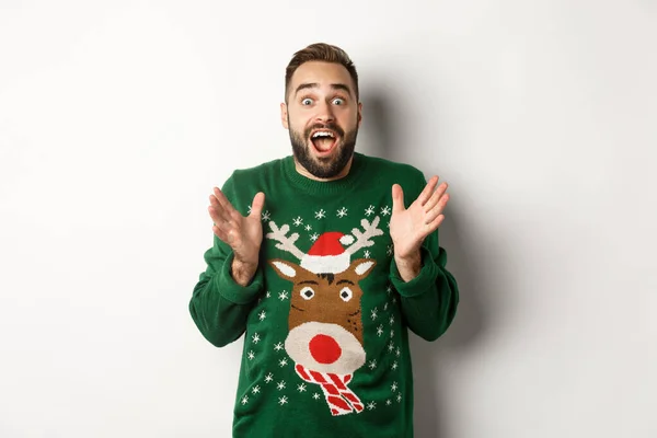 Año nuevo celebración y vacaciones de invierno concepto. Chico barbudo feliz y sorprendido mirando sorprendido, cogiendo algo, de pie en divertido suéter de Navidad, fondo blanco —  Fotos de Stock