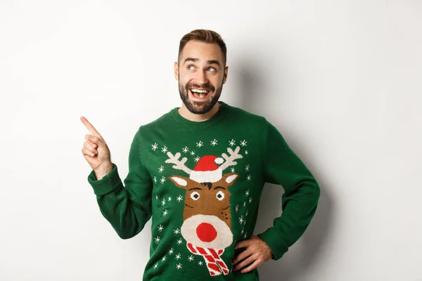 Nový rok oslavy a zimní prázdniny koncept. Zasněný vousatý muž v zeleném vánočním svetru, ukazuje na levý horní roh a pobaveně se usmívá, bílé pozadí — Stock fotografie