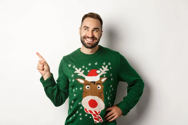 Nový rok oslavy a zimní prázdniny koncept. Hezký usmívající se muž v zeleném vánočním svetru, ukazuje a dívá se na levý horní roh obchodu s logem, bílé pozadí — Stock fotografie