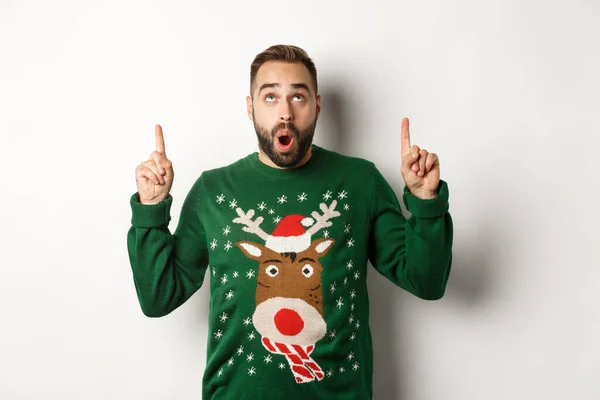 Зимові свята та Різдво. Дивовижний красивий чоловік у новорічному светрі, вказуючи пальці вгору, показуючи логотип, стоїть на білому тлі — стокове фото