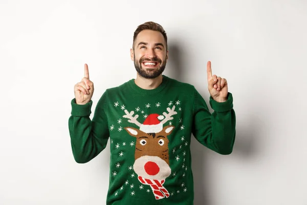 Wintervakantie en kerst. Gelukkig lachende man in grappige trui wijzende vingers omhoog, tonen reclame, staande over witte achtergrond — Stockfoto