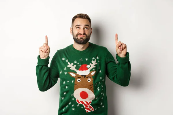 Vacanze invernali e Natale. Ragazzo scontento guardando dubbia spazio copia, puntando il dito verso l'alto e smorfia, in piedi su sfondo bianco — Foto Stock