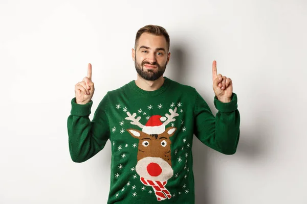 Зимові свята та Різдво. Ненасичений бородатий хлопець у смішному светрі вказує пальці вгору, показуючи щось неприємне, біле тло — стокове фото