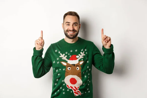Zimní prázdniny a Vánoce. Hezký vousatý muž v zeleném svetru, ukazuje prsty nahoru a usmívá se, ukazuje reklamu, bílé pozadí — Stock fotografie