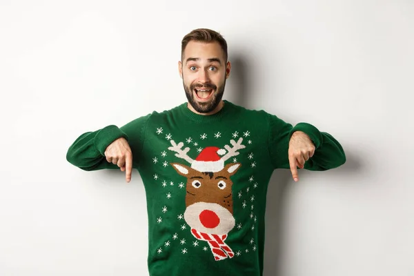 Kış tatili ve Noel. Yeşil kazaklı, heyecanlı sakallı adam, parmakları aşağıda ve şaşkın görünüyor, beyaz arka planda duruyor. — Stok fotoğraf