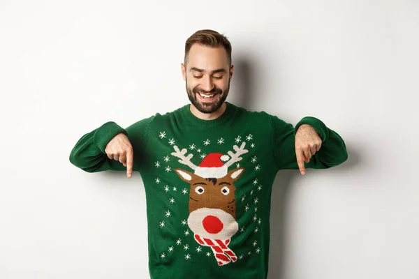 Vacaciones de invierno y Navidad. Hombre caucásico feliz en suéter verde, apuntando con los dedos hacia abajo y mirando satisfecho, de pie sobre fondo blanco —  Fotos de Stock