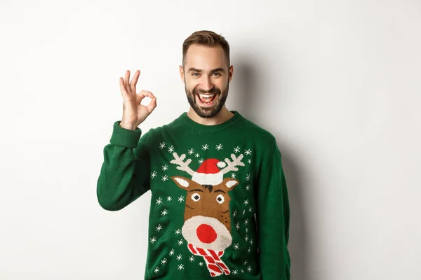 Vacaciones de invierno y Navidad. Hombre barbudo satisfecho en suéter verde, mostrando signo OK en aprobación, como algo bueno, de pie sobre fondo blanco —  Fotos de Stock