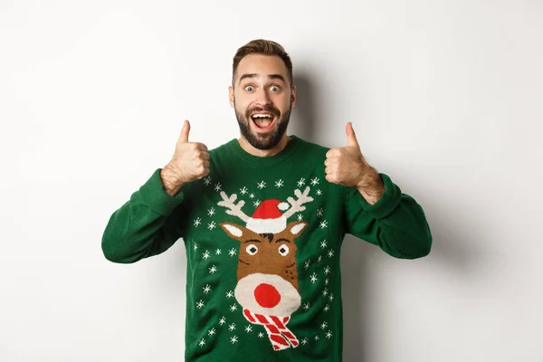 Navidad, fiestas y celebración. Tipo barbudo excitado en suéter verde mostrando pulgares hacia arriba, como algo increíble, de pie sobre fondo blanco — Foto de Stock