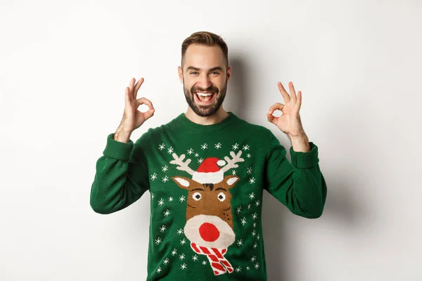 Navidad, fiestas y celebración. Tipo guapo satisfecho en suéter, mostrando signos de bien y sonriendo, alabar algo grande, de pie sobre fondo blanco — Foto de Stock