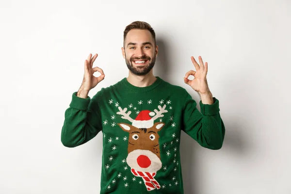 Χριστούγεννα, γιορτές και γιορτές. Ικανοποιημένος χαμογελαστός άνδρας με πράσινο πουλόβερ που δείχνει OK πινακίδες και γνέφει με έγκριση, συνιστώντας προϊόν, στέκεται πάνω από το λευκό φόντο — Φωτογραφία Αρχείου