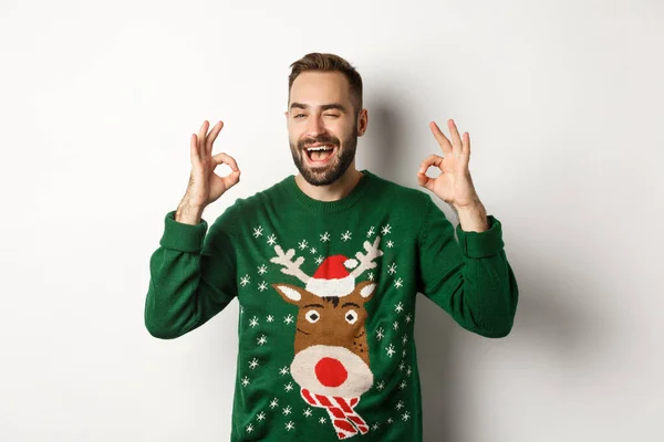 Natale, vacanze e feste. Felice giovane uomo in maglione verde, strizzando l'occhio e mostrando segni positivi, garantire la qualità, raccomandare qualcosa, sfondo bianco — Foto Stock