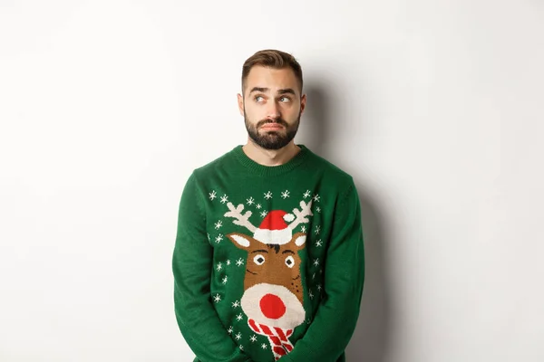 Capodanno, vacanze e feste. Uomo barbuto cupo in maglione, guardando indeciso e guardando da parte, in piedi su sfondo bianco — Foto Stock