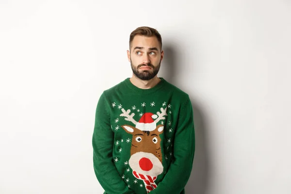 Nieuwjaar, feestdagen en feestdagen. Gloomy bebaarde man in trui, op zoek besluiteloos en opzij te kijken, staande tegen een witte achtergrond — Stockfoto