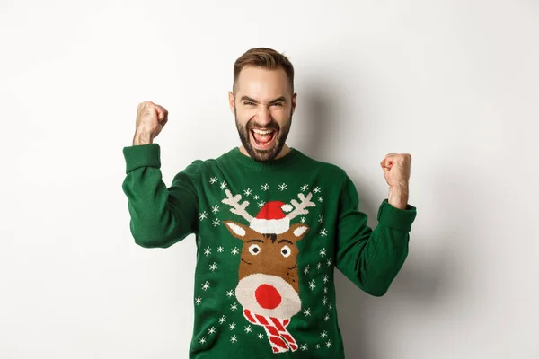 Año Nuevo, fiestas y celebración. Hombre barbudo excitado en suéter de Navidad, haciendo bombas de puño y gritando de alegría, regocijo y triunfo, fondo blanco — Foto de Stock