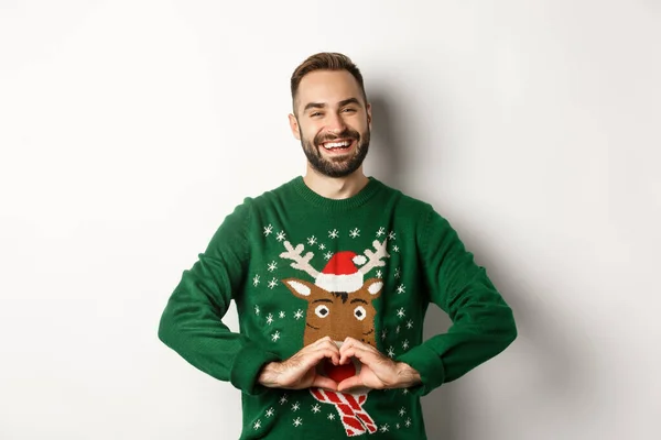 Новий рік, свята та святкування. Щасливий бородатий чоловік у різдвяному светрі показує знак серця, висловлює любов і турботу, стоїть над білим тлом — стокове фото