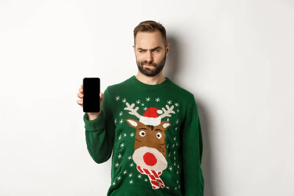 Neujahr, Feiertage und Feiern. Skeptischer Typ schaut zweifelnd auf den Bildschirm, während er es Ihnen zeigt, zeigt mobile App, steht über weißem Hintergrund — Stockfoto