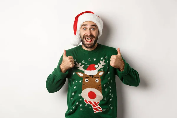 Vacaciones de Navidad, celebración y concepto de fiesta. Chico barbudo feliz en Santa sombrero mostrando pulgares hacia arriba en la aprobación, como algo impresionante, fondo blanco —  Fotos de Stock