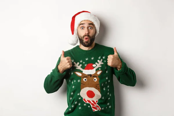 Vánoční svátky, oslavy a párty koncept. Úžasný mladý muž v Santa klobouk a svetr, ukazující palec nahoru v uznání, jako něco, bílé pozadí — Stock fotografie
