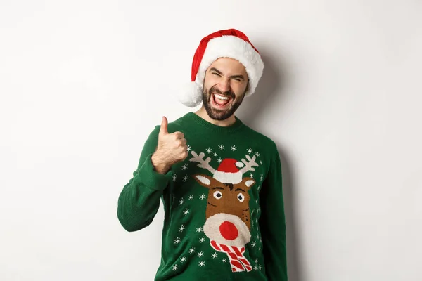 Christmas holidays, celebration and party concept. Man enjoying New Year, showing thumb up in approval, wearing Santa hat, white background — Stock Photo, Image