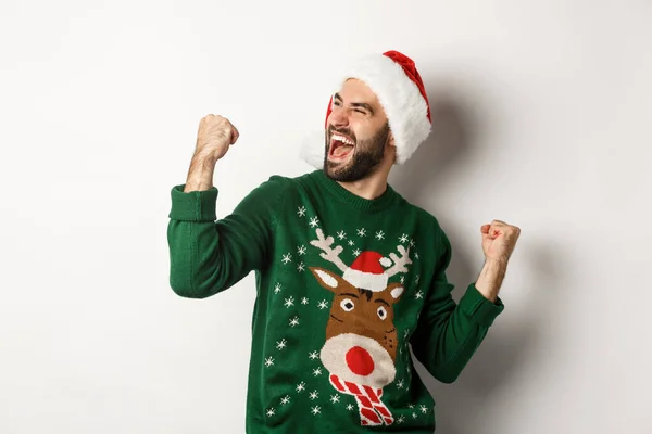 Feriados de Natal, celebração e conceito de festa. Cara feliz em chapéu de Papai Noel e suéter, fazendo bombas punho e alegria, triunfando, de pé sobre fundo branco — Fotografia de Stock