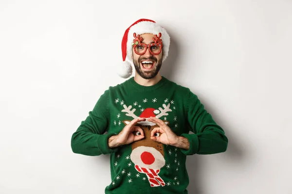 크리스마스 연휴, 축하 할 일. 산타 모자쓴 행복 한 남자와 당상 안경을 쓴 사람이 하얀 배경 위에 서서 웃긴 스웨터를 비웃고 있습니다. — 스톡 사진