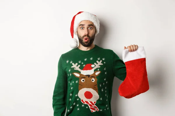 Koncept vánočních a zimních prázdnin. Happy man got a gift in Christmas sock, looking excited, standing in Santa hat against white background — Stock fotografie