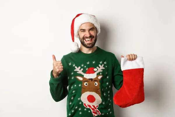 Xmas ve kış tatili konsepti. Noel Baba şapkalı mutlu ve memnun bir adam, Noel çorabında hediyeyi seviyor, başparmağını kaldırıyor, beyaz arka planda duruyor. — Stok fotoğraf