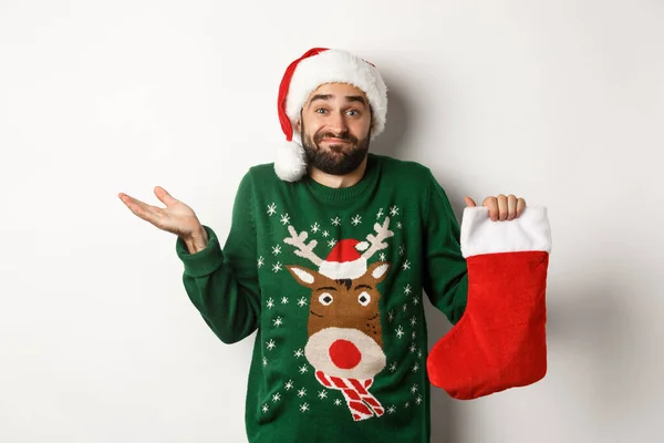 Koncept vánočních a zimních prázdnin. Zmatený chlap v klobouku Santa drží vánoční ponožku a pokrčení rameny, stojí bezradný proti bílému pozadí — Stock fotografie