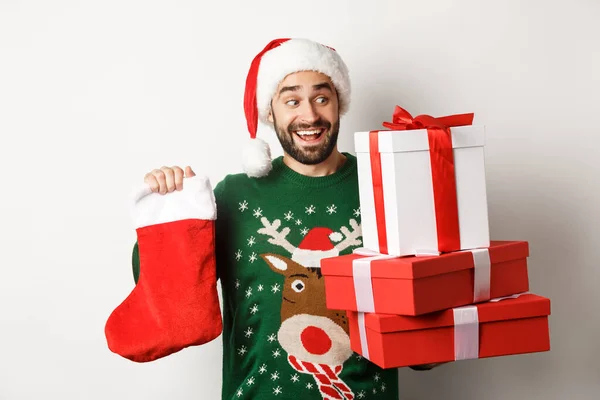 Koncept vánočních a zimních prázdnin. Nadšený muž drží vánoční ponožky a dárkové krabice, slaví Nový rok, stojí nad bílým pozadím — Stock fotografie