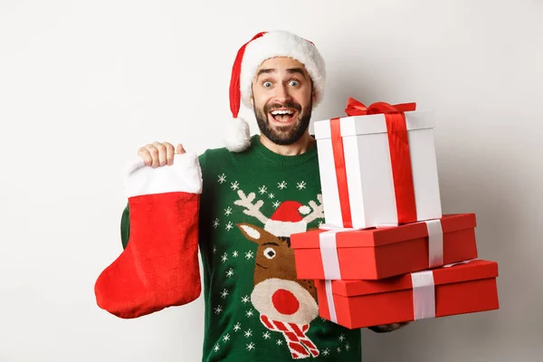 Koncept vánočních a zimních prázdnin. vzrušený muž drží vánoční ponožky a dárkové krabice, slaví Nový rok, přináší dárky pod stromeček, stojí nad bílým pozadím — Stock fotografie