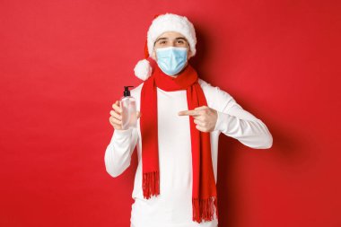 Covid-19, noel ve salgın sırasında tatil kavramı. Noel Baba şapkası ve tıbbi maskesiyle yakışıklı, el dezenfektanı tavsiye eden, kırmızı arka planda duran.