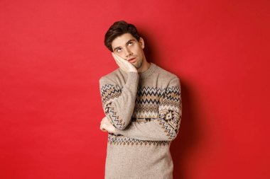 Yılbaşı süveteri giymiş, gözlerini devirmiş, eline yaslanmış, kırmızı arka planda duran, hoşnutsuzluğunu ve asabiyetini ifade eden sinirli genç bir adam görüntüsü.