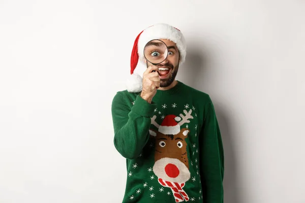 Концепція Різдва та свят. Смішний бородатий хлопець у капелюсі Санта, дивлячись через збільшувальне скло з подивом, знайшов щось, стоячи над білим тлом — стокове фото