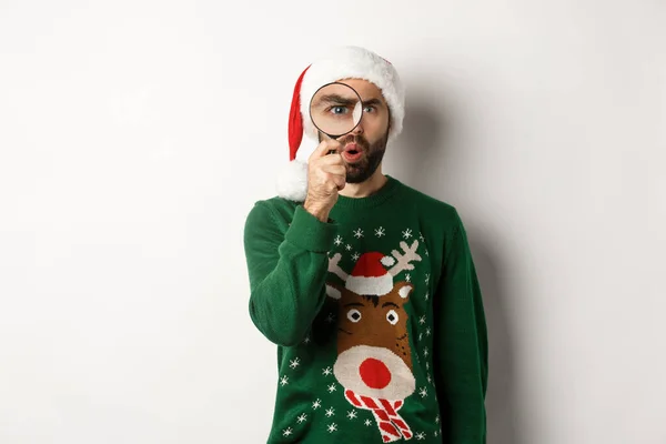 Vánoční a sváteční koncept. Zvědavý chlap dívá na něco přes lupu, stojící v Santa klobouk s vánočním svetrem, stojící nad bílým pozadím — Stock fotografie