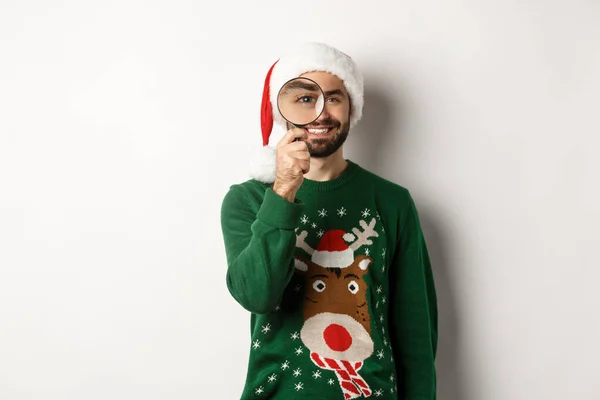 Noel ve tatil konsepti. Noel Baba şapkalı ve xmas kazaklı mutlu sakallı adam büyüteçle bakıyor ve gülümsüyor, beyaz arka planda duruyor. — Stok fotoğraf