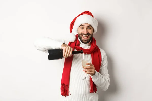 Weihnachtsfeier und Ferienkonzept. Glücklicher bärtiger Mann mit Weihnachtsmütze und Schreckschusspistole, übergießt sich mit Champagner und lächelt, feiert Neujahr, steht vor weißem Hintergrund — Stockfoto