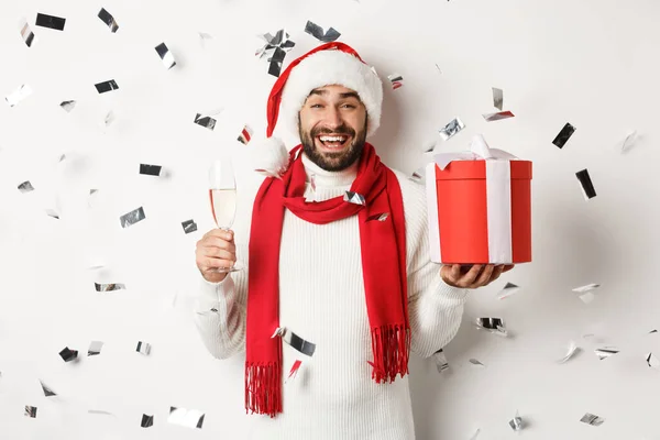 Festa de Natal e conceito feriados. Cara feliz comemorando o Ano Novo, segurando presente e vidro, bebendo champanhe e rindo, confete voando ao redor, fundo branco — Fotografia de Stock