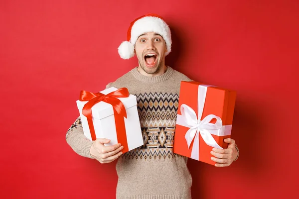 Wizerunek wesołego atrakcyjnego faceta trzymającego prezenty świąteczne, stojącego w czapce Mikołaja i zimowym swetrze, uśmiechniętego zdumionego, stojącego nad czerwonym tłem — Zdjęcie stockowe