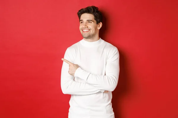 Begrepet vinterferie, jul og livsstil. Kjekk voksen mann i hvit genser, ser ut og peker til venstre med et gledelig smil, viser reklame for nyttår, rød bakgrunn – stockfoto