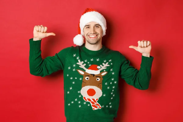 겨울 휴가, 크리스마스, 생활 양식의 개념. 산타 모자와 스웨터를 입고 빨간 배경 위에 서서 만족 스러운 얼굴로 자신을 가리키는 행복 한 매력적 인 남자 — 스톡 사진