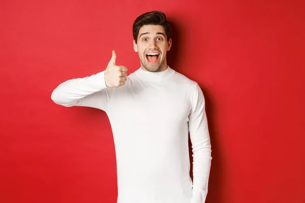 Concept of winter holidays, christmas and lifestyle. Excited good-looking man in white sweater, like good product, showing thumb-up in approval and smiling amazed, red background — Stock Photo, Image